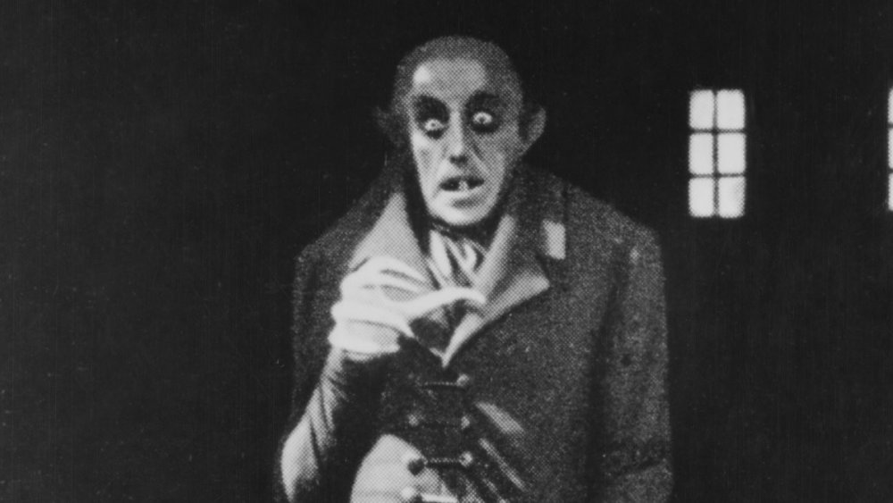 Black and white image of a creepy figure with large eyes and sharp teeth in a dark coat, set against a dim background with two small windows.