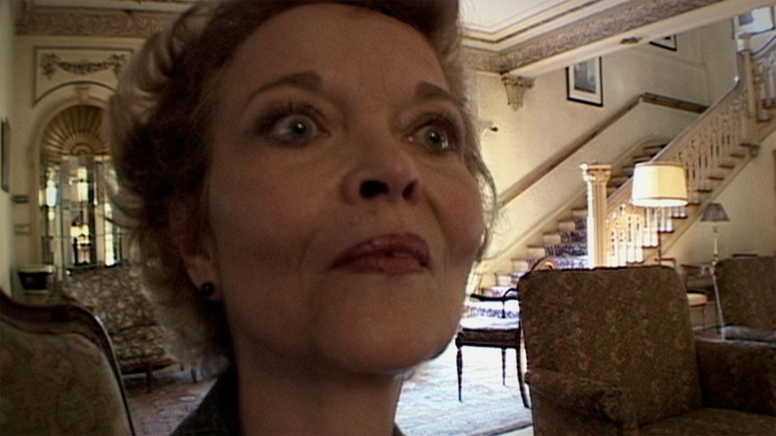 Close-up of an older woman in a vintage-style room with a grand staircase.