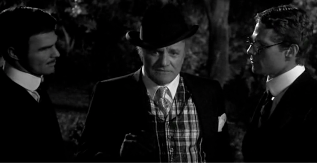 A still from the climax of Peter Bogdanovich's film 'Nickelodeon'. An older man in a hat looks toward the camera, in deep thought, and two men, at either side of him, look on listening.